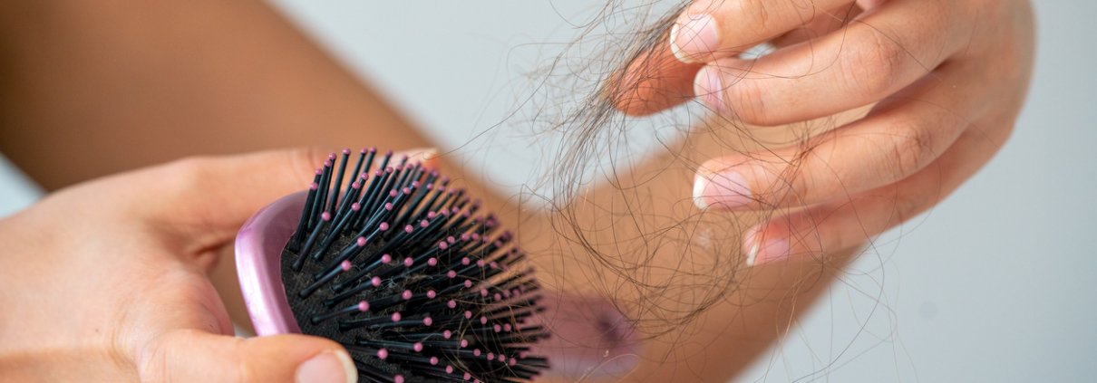 A woman holding a light pink hair brush and picking her hair out of it.